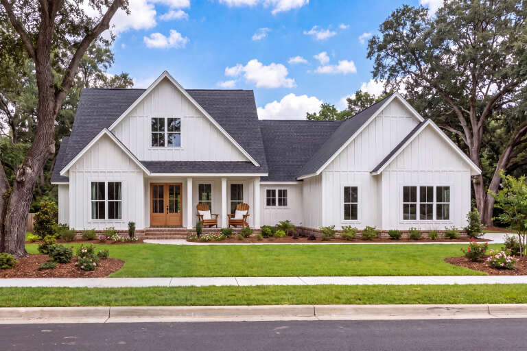 White Modern Farmhouse, board-and-batten siding, dark roof, wooden front door, porch chairs, and lush green landscaping. House Plan # 041-00351 Digital Rendering by America's Best House Plans, Inc.