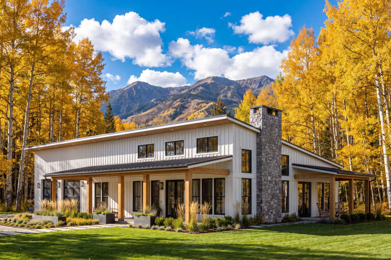 Angled front view of a white Barndominium house, stone chimney, wood porch posts, black windows, and mountain backdrop. House Plan # 041-00388