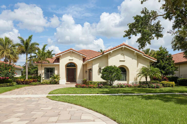 Front view of a beige house in Mediterranean style, red tile roof, arched entry, and lush tropical landscaping. House Plan # 8436-00137