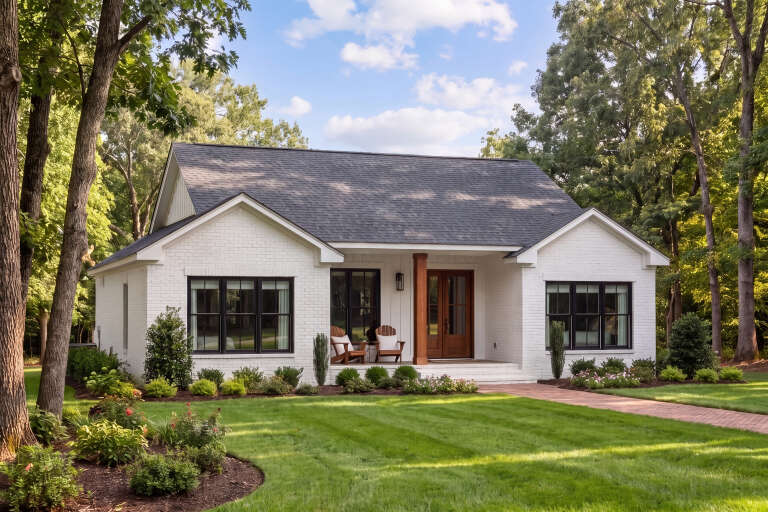 White brick cottage, Angled Front Elevation, black window frames, wooden front door, porch chairs, brick walkway, trees. House Plan # 041-00453 Digital Rendering by America's Best House Plans, Inc.
