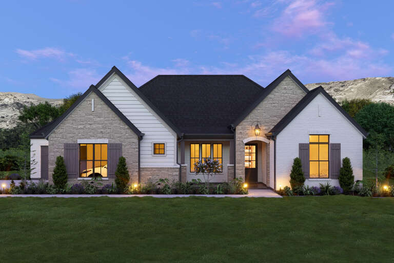 White French Country, Front Elevation at dusk, stone and brick siding, wood shutters, lit windows, and lush landscaping. House Plan # 3571-00045 Digital Rendering by America's Best House Plans, Inc.