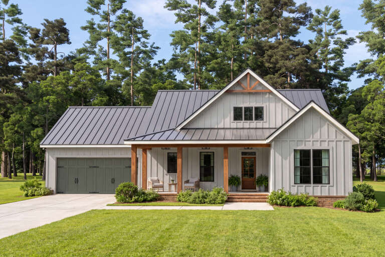 Front view of a grey house, board-and-batten siding, metal roof, wood porch posts, green garage door, and lush landscape. House Plan # 009-00539