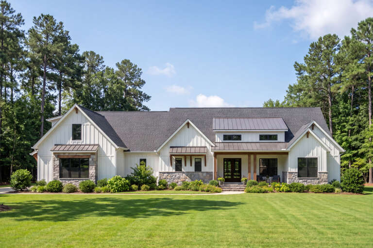 Front view of a white house in Modern Farmhouse style, stone base, black windows, metal roof accents, and lush landscape. House Plan # 009-00542