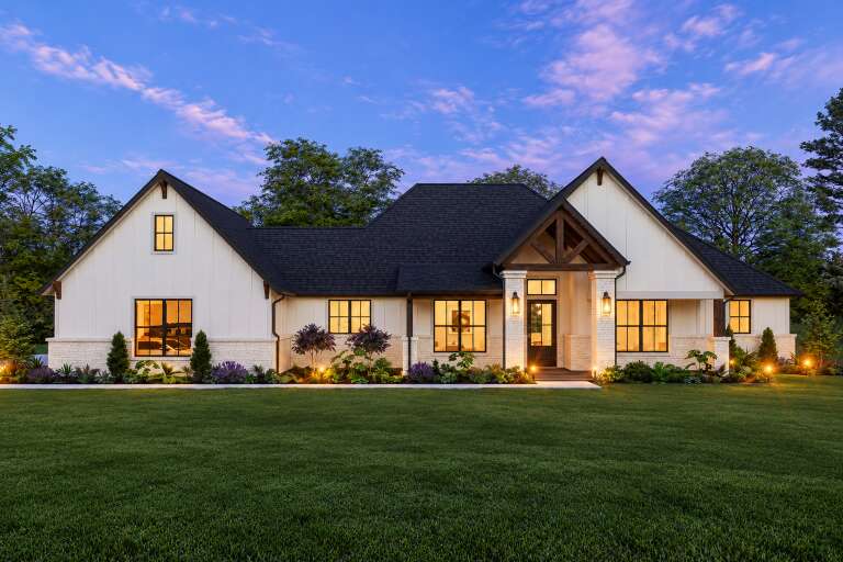 Front view of a white house in Modern Farmhouse style, board-and-batten siding, stone base, dark roof, and lit porch. House Plan # 3571-00047