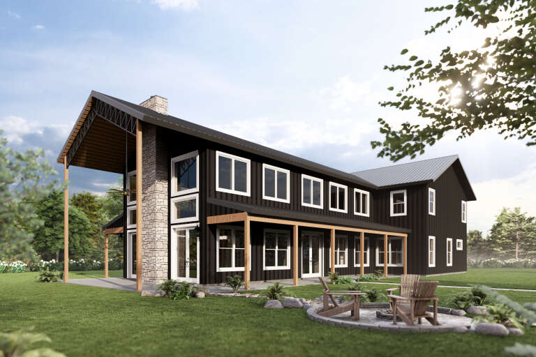 Angled rear view of a black house in Barndominium style, stone chimney, metal roof, covered porch, and fire pit area. House Plan # 3822-00063