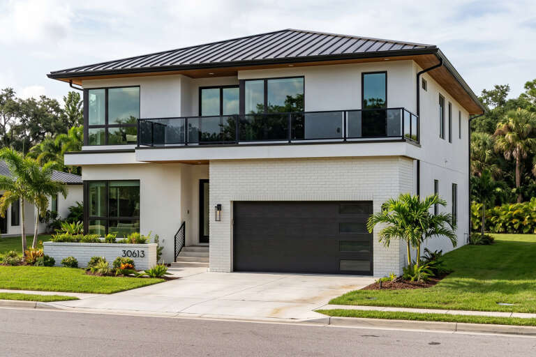Angled front view of a white house in Modern style, brick accents, metal roof, glass balcony, and dark garage door. House Plan # 963-01348