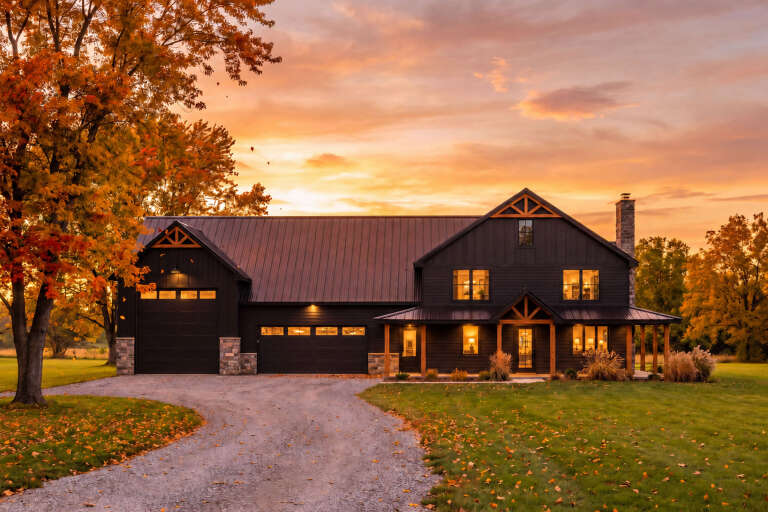Front view of a black Barndominium house, metal roof, stone accents, timber trusses, and an illuminated covered porch. House Plan # 402-01940