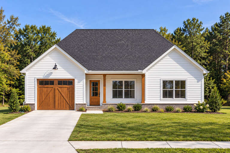 Front view of a white house, wood garage door, brick base, dark roof, wood porch posts, and manicured landscaping. House Plan # 7568-00072
