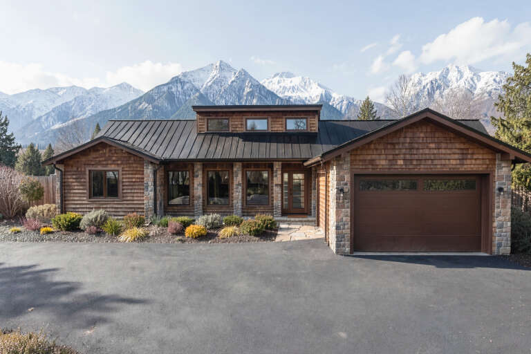 Front view of a brown house, cedar shake siding, stone accents, black metal roof, mountain backdrop, paved driveway. House Plan # 4440-00029