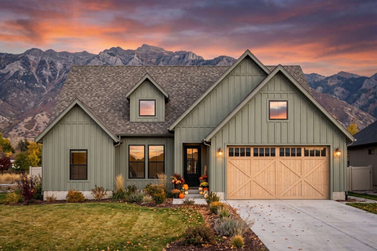 Front view of a sage green house in Modern Farmhouse style, board-and-batten siding, wood garage door, mountain backdrop. House Plan # 7983-00224